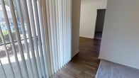 This single-level space offers a clean, functional layout designed with simplicity and practicality in mind. The interior is finished with warm hardwood-style flooring that runs throughout the main areas, creating a cohesive and inviting feel underfoot. Neutral white walls provide a bright, versatile backdrop that complements a variety of furnishing styles and décor choices.

A hallway connects the key areas of the space, offering a clear sense of flow and easy navigation between rooms. Vertical blinds line the windows, providing effective light control while allowing natural light to softly illuminate the interior when open. The overall color palette is understated and modern, keeping the atmosphere light and airy.

The rooms visible through the hallway maintain the same consistent flooring and wall finishes, contributing to a unified interior aesthetic throughout. The space presents a practical and well-maintained environment, suitable for a range of uses. Its straightforward design ensures ease of movement and a comfortable, welcoming ambiance that is both functional and adaptable to personal customization.