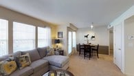This single-floor residence offers a seamlessly connected living and dining layout designed for comfortable everyday living. The living area is anchored by plush, neutral-toned carpeting and features a generously sized sectional sofa complemented by decorative accent pillows, creating an inviting and relaxed atmosphere. Large windows dressed with white horizontal blinds flood the space with abundant natural light, enhancing the warm, neutral tones of the walls.

The dining area transitions naturally from the living space and is defined by a two-tone wall treatment, with a darker wainscoting panel below and a soft beige upper wall, adding a touch of architectural character. A round dark-wood dining table surrounded by matching chairs sits beneath a track lighting fixture, providing focused illumination for meals and gatherings. Wall art adds a personalized, homey touch to the dining nook.

Throughout the space, the open floor plan promotes an airy sense of connectivity, while the consistent carpeting and cohesive color palette tie the rooms together harmoniously. The combination of ample natural light, thoughtful design details, and comfortable furnishings makes this a welcoming and functional living environment.