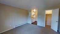 This single-floor unit offers a straightforward, functional layout designed to maximize everyday livability. The living area is finished with wall-to-wall carpet in a neutral gray tone, providing a comfortable underfoot feel, while the textured popcorn ceiling adds a classic touch overhead. Warm beige walls create a neutral backdrop throughout the space. A baseboard heater runs along the far wall of the living area, ensuring efficient warmth during cooler months.

The kitchen is positioned just off the main living space and is distinguished by its dark hardwood-style flooring, creating a clear visual separation between the two areas. White cabinetry lines the upper and lower walls, offering ample storage, while white appliances — including a refrigerator, stove, and sink with a window above — complete the setup. Overhead lighting illuminates the kitchen area effectively.

To the right of the living space, a doorway leads to what appears to be additional rooms, with a closet visible nearby, suggesting practical storage options. The overall ambiance is clean, neutral, and move-in ready.