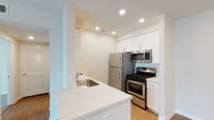 This single-floor residence showcases a clean, contemporary design with a bright and airy aesthetic throughout. The kitchen is a standout feature, thoughtfully designed with crisp white shaker-style cabinetry that extends to the ceiling, offering generous storage. A suite of stainless steel appliances — including a gas range, over-the-range microwave, and a full-size refrigerator — provides a modern, functional cooking environment. The kitchen peninsula, finished with a sleek white countertop, incorporates a single-basin sink and creates a natural divide between the kitchen and the adjacent living area, making it ideal for casual dining or entertaining. Light wood-tone flooring runs underfoot, adding warmth and continuity to the space, while recessed lighting in the ceiling ensures the area is well-lit at any time of day. The walls are finished in soft, neutral tones that complement the white cabinetry and bright countertops, creating a cohesive and inviting atmosphere. The overall layout is efficient and well-organized, maximizing the available space while maintaining an open, uncluttered feel throughout the home.