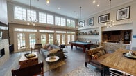 Located at 3750 E New York St in Aurora, IL, this single-floor community clubhouse is designed to offer residents a well-appointed gathering and recreation space. The centerpiece of the clubhouse is a large, open-concept lounge area distinguished by soaring ceilings, crown molding, and an abundance of natural light streaming through a striking wall of tall, multi-pane windows and French doors that overlook the outdoor pool area. The lounge is furnished with plush sofas, tufted leather seating, and layered area rugs, creating a warm and inviting atmosphere. A billiards table anchors the recreational zone, complemented by a built-in banquette and display shelving adorned with curated décor and Chicago-themed artwork. A linear gas fireplace set into a herringbone wood-panel accent wall adds a cozy focal point, with a wall-mounted flat-screen TV above. Adjacent to the fireplace, a dedicated workspace features built-in open shelving stocked with books and decorative objects, paired with leather rolling chairs and desktop workstations. The entry area transitions seamlessly into the main lounge, showcasing large-format tile flooring throughout. A communal dining table with striped seating and a wet bar with mosaic tile backsplash round out the space, making it ideal for both casual gatherings and productive work sessions.