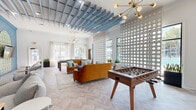 This stylish communal lounge space is a showcase of bold, mid-century modern design. The open-plan layout is unified by striking herringbone-patterned hardwood flooring and soaring ceilings adorned with distinctive powder-blue slatted ceiling panels that add architectural depth and a playful sense of dimension. Sputnik-style chandeliers in brushed gold serve as elegant focal points throughout, casting warm ambient light across the space.

A standout decorative partition wall featuring a repeating circular cut-out pattern divides the room while allowing natural light to filter through from large perimeter windows, offering views of the surrounding outdoor landscape. The space is furnished with rich amber-toned velvet sofas and accent chairs, creating vibrant seating areas ideal for socializing and relaxation.

A dedicated gaming area includes a full-size foosball table and a shuffleboard table, reinforcing the space's recreational purpose. A deep teal accent wall hosts three arched niches framed with bold geometric-patterned wallpaper, each housing a mounted flat-screen television — a visually striking entertainment feature. A sculptural wave-form ceiling detail crowns this lounge zone. Lush potted greenery is placed throughout, adding organic warmth and contrast to the contemporary palette.