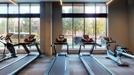 This fitness center is thoughtfully designed to accommodate a full range of exercise needs within a single, open-plan level. The space is divided into two primary zones: a cardio area and a strength training area, each finished with distinct flooring to delineate function. The cardio zone is anchored by a row of Precor treadmills and elliptical machines positioned directly in front of expansive floor-to-ceiling windows. These large, grid-paned windows flood the area with natural light and offer views of surrounding greenery and the urban streetscape beyond, creating an energizing atmosphere during workouts. Structural columns punctuate the space, adding an architectural rhythm without interrupting the open feel. The strength training area features a selection of cable machines, a multi-station weight system, adjustable benches, and a dumbbell rack. A teal accent wall paired with large wall-mounted mirrors visually expands this zone while allowing users to monitor their form. Linear LED lighting runs across the ceiling, providing consistent, bright illumination throughout. A water fountain station is conveniently integrated along the wall. The overall design balances a clean, contemporary aesthetic with practical functionality.