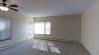 This single-floor space presents a straightforward, functional layout centered around a spacious main living area. The room features neutral cream-colored walls and a smooth white ceiling, creating a clean, understated aesthetic that serves as a versatile backdrop for a variety of interior styles.

A prominent window fitted with vertical blinds allows generous amounts of natural light to filter into the room, casting distinct light patterns across the carpeted floor throughout the day. The window can be opened to allow airflow, adding a practical element to the bright atmosphere.

The flooring throughout the main living area consists of wall-to-wall carpeting in a neutral tone, contributing to a quiet and comfortable underfoot feel. Baseboard heating units run along the base of the walls, providing an efficient and unobtrusive heating solution during colder months.

The overall ambiance of the space is calm and minimal, with clean lines and an open feel that allows for flexible furniture arrangement. The lack of clutter and the abundance of natural light make the room feel airy and welcoming. The single-floor design ensures easy accessibility and a cohesive flow throughout the space.