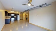 This single-floor unit showcases an open-concept layout that seamlessly integrates the living area and kitchen into one continuous space. The polished concrete flooring runs throughout, lending a clean, industrial aesthetic that is both durable and visually distinctive. An exposed metal duct runs along the ceiling, complementing the utilitarian design, while a ceiling fan with a built-in light fixture provides both airflow and ambient lighting to the main living area.

The kitchen, situated toward one end of the space, is equipped with stainless steel appliances, including a refrigerator, gas range, and overhead microwave. Dark cabinetry provides strong contrast against the light-toned walls, and pendant lighting adds a warm glow to the workspace.

Toward the rear of the unit, a set of doors leads to additional spaces, including what appears to be a bathroom with visible cabinetry and mirror, as well as an in-unit laundry area with stacked washer and dryer units. The light yellow baseboard trim and cream-colored walls contribute a subtle warmth that balances the space's industrial undertones, creating a practical yet characterful interior.