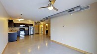 This single-floor unit showcases an open-concept layout that seamlessly integrates the living area and kitchen into one continuous space. The polished concrete flooring runs throughout, lending a clean, industrial aesthetic that is both durable and visually distinctive. An exposed metal duct runs along the ceiling, complementing the utilitarian design, while a ceiling fan with a built-in light fixture provides both airflow and ambient lighting to the main living area.

The kitchen, situated toward one end of the space, is equipped with stainless steel appliances, including a refrigerator, gas range, and overhead microwave. Dark cabinetry provides strong contrast against the light-toned walls, and pendant lighting adds a warm glow to the workspace.

Toward the rear of the unit, a set of doors leads to additional spaces, including what appears to be a bathroom with visible cabinetry and mirror, as well as an in-unit laundry area with stacked washer and dryer units. The light yellow baseboard trim and cream-colored walls contribute a subtle warmth that balances the space's industrial undertones, creating a practical yet characterful interior.