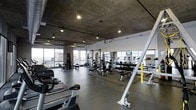 This modern fitness center offers a comprehensive workout environment designed to meet a wide range of training needs. The space is defined by its industrial-inspired aesthetic, featuring exposed concrete ceilings with recessed and track lighting that creates a focused, energizing atmosphere. Large floor-to-ceiling windows line the perimeter, flooding the space with natural light and offering expansive views of the surrounding cityscape. The open layout is thoughtfully organized into distinct training zones. A dedicated strength training area houses a full rack of dumbbells, multiple weight benches, and a selection of cable and multi-function machines, providing everything needed for resistance training. Cardio equipment, including stationary bikes, treadmills, and rowing machines, is positioned to take advantage of the natural light and views. Rubberized flooring in the free weights section provides safety and durability, while polished concrete floors extend throughout the rest of the space. Mirrored walls in key areas enhance the sense of space and allow for proper form checks during exercise. The overall ambiance is sleek, functional, and motivating — a well-appointed facility suited for a serious fitness regimen.