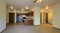 This single-floor residence offers a practical and well-organized layout designed for comfortable everyday living. The heart of the home is an open-concept kitchen and living area that flows seamlessly together, creating a spacious and inviting atmosphere. The kitchen is equipped with rich, dark wood cabinetry that provides ample storage, a black refrigerator, a black range, and a sink set into a light-toned countertop. A peninsula-style breakfast bar extends from the cabinetry, defining the kitchen space while maintaining an open connection to the carpeted living area. Recessed and flush-mount ceiling lighting illuminates the kitchen, while a ceiling fan with a light fixture keeps the living area bright and comfortable. The neutral-toned carpet throughout the living space adds warmth underfoot. A structural column adds architectural interest to the open floor plan, subtly dividing functional zones without closing off the space. From the main living area, doorways lead to additional rooms, including what appears to be bedrooms, offering privacy while maintaining easy access to the shared living spaces. The overall ambiance is clean, functional, and move-in ready.