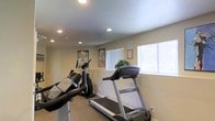 This single-floor fitness room offers a functional workout space designed to support cardiovascular exercise. The room features carpeted flooring and smooth, neutral-toned walls that create a clean, understated environment. Recessed ceiling lights provide consistent, even illumination throughout the space, complemented by natural light streaming in through two windows fitted with vertical blinds. The windows also offer views of the surrounding exterior, adding an open feel to the room. Equipment includes a treadmill, an elliptical trainer, and a stationary bike, positioned to allow adequate space for use. Informational fitness posters and a wall clock are mounted along the walls, adding practical and motivational elements to the space. A small potted plant near the windows introduces a touch of greenery. The textured ceiling and neutral color palette contribute to a straightforward, utilitarian atmosphere suited for regular exercise. Overall, the space is efficiently organized to maximize its footprint while providing residents or users with essential cardio fitness options in a well-lit, accessible setting.