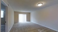 This single-floor space presents a clean and functional layout designed with simplicity in mind. The main living area is finished with wall-to-wall carpeting in a neutral beige tone, providing a soft and comfortable underfoot feel. Smooth, white baseboards line the perimeter of the room, adding a crisp contrast against the light beige walls. The textured ceiling features a classic popcorn finish and is equipped with a flush-mount ceiling light fixture, ensuring consistent overhead illumination throughout the space. A prominent sliding glass door fitted with vertical blinds serves as the focal point of the main room, allowing an abundance of natural light to filter in while offering privacy control. The vertical blinds can be adjusted to regulate light levels as needed. The overall color palette remains understated and neutral throughout, creating a versatile backdrop well-suited for a variety of interior design styles. The single-floor layout offers ease of accessibility, and the open, unobstructed floor plan provides flexibility in how the space can be arranged and utilized.