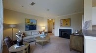 Located at 100 Harpers Crossing in Langhorne, Pennsylvania, this single-floor residence offers a well-appointed and comfortable living environment with thoughtful architectural details. The living room serves as a central gathering space, featuring a classic fireplace with a clean white surround and dark firebox that anchors the room as a focal point. Crown molding runs along the ceiling, adding a refined touch to the overall design. Neutral-toned carpeting and warm beige walls create a calm, inviting ambiance throughout. The space is illuminated by a combination of recessed ceiling lighting and soft accent lamps, contributing to a welcoming atmosphere. A ceiling-mounted speaker suggests an integrated audio system for added convenience. The layout flows naturally from the living area toward adjacent rooms, with a hallway visible in the background connecting to additional spaces within the home. Built-in ventilation is incorporated into the ceiling design, reflecting practical attention to comfort. The room comfortably accommodates seating arrangements and decorative furnishings, highlighting its versatility. Overall, this single-story home presents a clean, classic aesthetic with functional design elements suited for everyday living.