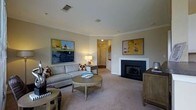 Located at 100 Harpers Crossing in Langhorne, Pennsylvania, this single-floor residence offers a well-appointed and comfortable living environment with thoughtful architectural details. The living room serves as a central gathering space, featuring a classic fireplace with a clean white surround and dark firebox that anchors the room as a focal point. Crown molding runs along the ceiling, adding a refined touch to the overall design. Neutral-toned carpeting and warm beige walls create a calm, inviting ambiance throughout. The space is illuminated by a combination of recessed ceiling lighting and soft accent lamps, contributing to a welcoming atmosphere. A ceiling-mounted speaker suggests an integrated audio system for added convenience. The layout flows naturally from the living area toward adjacent rooms, with a hallway visible in the background connecting to additional spaces within the home. Built-in ventilation is incorporated into the ceiling design, reflecting practical attention to comfort. The room comfortably accommodates seating arrangements and decorative furnishings, highlighting its versatility. Overall, this single-story home presents a clean, classic aesthetic with functional design elements suited for everyday living.