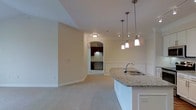 This single-floor residence offers a thoughtfully designed open-concept layout that seamlessly connects the living and kitchen areas. The kitchen is highlighted by a granite-topped peninsula with an undermount sink, served by a trio of sleek pendant lights that provide both task lighting and visual elegance. Rich dark-toned cabinetry, visible through a graceful arched passageway, adds warmth and contrast to the otherwise light and neutral color palette throughout the space.

The living area features plush wall-to-wall carpeting in a soft neutral tone, creating a comfortable and inviting atmosphere. Crown molding and classic wainscoting panels add a refined, traditional character to the walls, while recessed lighting and flush-mount ceiling fixtures ensure the space is well-lit throughout.

The arched entryway serves as a distinctive architectural focal point, elegantly framing the transition between the living space and the kitchen beyond. Smooth, cream-toned walls and understated finishes contribute to a clean, cohesive aesthetic that feels both functional and welcoming. Overall, this residence combines classic architectural detailing with modern conveniences to create a polished and comfortable living environment.