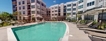 Located at 6 Becker Farm Rd in Roseland, NJ, this outdoor amenity space centers around a large, resort-style swimming pool with crystal-clear turquoise water. The pool features a gently sloping shallow entry area with in-water lounge chairs, making it ideal for relaxation, as well as stainless steel entry railings for convenient access. The expansive pool deck is finished in light-toned pavers and is lined with numerous slatted chaise lounge chairs arranged throughout the space. Multiple market umbrellas provide shaded seating areas, while pergola structures with translucent privacy screens offer additional sheltered lounging spots with side tables. Lush green lawn areas and manicured landscaping accent the perimeter of the deck. The pool area is enclosed by a black iron safety fence and is set within a courtyard formed by the surrounding multi-story residential building, which features a contemporary architectural design with large windows, private balconies, and a neutral gray and white exterior. The entire space is open to abundant natural light and blue skies, creating a vibrant and welcoming outdoor retreat for residents.