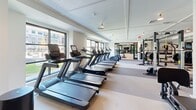 This expansive fitness center is thoughtfully designed to accommodate a full range of workout needs within a clean, modern aesthetic. The space is organized into distinct functional zones, each outfitted with professional-grade equipment and finished with bright white walls, coffered ceilings, and light-toned flooring throughout.

The cardio zone lines the perimeter walls, featuring multiple treadmills and cardio machines positioned alongside large windows that flood the area with natural light and offer views of the surrounding exterior. The strength training area is generously equipped with a cable machine, leg press, power rack, adjustable benches, kettlebells, dumbbells, a punching bag, and a rowing machine, catering to a wide variety of training styles.

A dedicated studio room provides a versatile space for yoga, stretching, or group fitness. This room features warm hardwood-style flooring, floor-to-ceiling mirrors, and an organized array of accessories including resistance tools, stability balls, and exercise mats.

Near the entrance, a welcoming reception area includes open shelving, a beverage cooler, and sleek glass-panel doors that frame views into the main gym floor. Wall-mounted fans, accent lighting, and motivational artwork complete the polished, professional atmosphere of this comprehensive fitness facility.