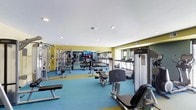 This fitness center is designed to accommodate a comprehensive workout experience within a single-floor layout. The space is finished with bold yellow accent walls and blue flooring, creating an energetic and motivating atmosphere. Recessed lighting throughout ensures the room is well-lit, complementing the natural light that enters through windows along one side.

The cardio zone features a row of treadmills and elliptical machines positioned along a mirrored wall, allowing users to monitor their form during exercise. A wall-mounted flat-screen television is centrally placed, providing entertainment during workouts. The strength training area is well-stocked with a multi-station cable machine, a dumbbell rack, and various weight benches, offering a versatile selection for resistance training. Additional strength equipment, including a dedicated weight machine, is positioned toward the perimeter, maximizing the usable floor space.

The layout is thoughtfully organized to allow users to move between equipment without congestion. The mirrored wall enhances the sense of openness, making the space feel larger and more expansive than it might otherwise appear.