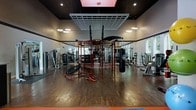 This single-floor fitness center offers a comprehensive workout environment designed to accommodate a wide variety of training styles and fitness goals. The space features rich hardwood-style flooring throughout, which contrasts sharply with the dark ceiling and walls, creating a focused and energizing atmosphere. At the heart of the gym stands a prominent multi-functional red power rack and cable rig system, which serves as the centerpiece for strength and functional training. Suspended from the ceiling above the rig is a grid panel system, ideal for suspension and resistance training accessories. A heavy punching bag hangs nearby, catering to combat fitness and conditioning workouts. Along the perimeter, a robust selection of selectorized strength machines and free weight stations are neatly arranged, supported by large mirrors that enhance the sense of space and allow users to monitor their form. Recessed and pendant lighting provides bright, even illumination across the floor, while a window on the far wall introduces natural light. Additional equipment including a mini trampoline and various specialty machines rounds out the facility's extensive offerings, making this a fully equipped space suited for all levels of fitness enthusiasts.