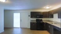 This 2-floor space features a kitchen equipped with dark espresso-toned cabinetry that extends to the upper walls, providing ample storage. The kitchen includes black appliances — a gas range and an over-the-range microwave — complemented by light granite-style countertops that contrast nicely with the dark cabinets. Light hardwood flooring runs throughout, adding warmth to the overall aesthetic. A textured ceiling adds subtle dimension to the space. Natural light enters through a large window fitted with blinds near the entry door, which features a deadbolt and an adjacent wall-mounted intercom or utility panel. The open layout of the kitchen area connects fluidly to the rest of the main floor, creating a functional and cohesive living environment. The neutral wall tones and consistent flooring contribute to a clean, modern feel throughout the space.