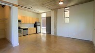 This stylish urban loft showcases an open-plan design that blends industrial character with modern functionality. The space is defined by its polished concrete floors and exposed concrete ceiling, which lend a raw, contemporary aesthetic throughout. Track lighting runs along the ceiling, providing flexible and functional illumination across the generous open living area.

The kitchen is seamlessly integrated into the main living space and features light wood cabinetry, a subway tile backsplash, stainless steel appliances including a refrigerator, dishwasher, and range, as well as a practical sink setup — making it both stylish and highly functional.

Natural light filters into the space through tall windows fitted with blinds, including a prominent floor-to-ceiling window flanking the entrance door, which draws in ample daylight and adds a sense of openness and volume to the interior. Additional windows on the adjacent wall further brighten the space.

The expansive open floor plan offers tremendous flexibility for furniture arrangement and customization, making it well-suited for a variety of uses. Exposed structural concrete columns punctuate the space, reinforcing the industrial loft character that defines this unique and versatile single-floor property.