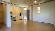 This stylish urban loft showcases an open-plan design that blends industrial character with modern functionality. The space is defined by its polished concrete floors and exposed concrete ceiling, which lend a raw, contemporary aesthetic throughout. Track lighting runs along the ceiling, providing flexible and functional illumination across the generous open living area.

The kitchen is seamlessly integrated into the main living space and features light wood cabinetry, a subway tile backsplash, stainless steel appliances including a refrigerator, dishwasher, and range, as well as a practical sink setup — making it both stylish and highly functional.

Natural light filters into the space through tall windows fitted with blinds, including a prominent floor-to-ceiling window flanking the entrance door, which draws in ample daylight and adds a sense of openness and volume to the interior. Additional windows on the adjacent wall further brighten the space.

The expansive open floor plan offers tremendous flexibility for furniture arrangement and customization, making it well-suited for a variety of uses. Exposed structural concrete columns punctuate the space, reinforcing the industrial loft character that defines this unique and versatile single-floor property.