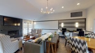 This thoughtfully designed single-floor communal space blends contemporary style with comfortable, functional living. The expansive open-plan lounge area serves as the heart of the space, anchored by a dramatic floor-to-ceiling dark stone fireplace with a wall-mounted television above. Multiple seating arrangements — including plush sofas, upholstered wingback chairs, and patterned accent chairs — invite relaxed gathering and conversation. A striking sputnik-style chandelier hangs overhead, complementing the vaulted ceiling and recessed lighting that together create a bright, welcoming atmosphere. Rich wood-look flooring flows throughout, tying the various zones together seamlessly. A dining area with a round wooden table and bold chevron-patterned chairs sits adjacent to a dedicated workstation nook, framed by a distinctive horizontal-striped accent wall. Natural light pours in generously through large French doors and oversized windows that offer views of the surrounding outdoors. The fully equipped kitchen is outfitted with crisp white shaker-style cabinetry, stainless steel appliances including a refrigerator, range, and microwave, white countertops, a glass tile backsplash, and an industrial-style dark wood island. The overall ambiance is modern yet inviting, balancing communal functionality with refined design details throughout.