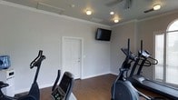 This single-floor fitness center offers a practical and well-maintained space designed for cardio-focused workouts. The room features smooth, light gray walls accented by crisp white crown molding, lending a polished finish to the functional environment. Hardwood-style flooring runs throughout, providing a durable and easy-to-clean surface suited for high-traffic use.

The space is equipped with multiple cardio machines, including ellipticals and a stationary bike, positioned to allow comfortable use and movement around the room. A wall-mounted flat-screen television is centrally placed, allowing users to watch content while exercising. Overhead, a combination of flush-mount and recessed lighting provides even, functional illumination throughout the room.

A paneled interior door provides access to an adjacent area, contributing to the overall flow of the facility. The HVAC system is visible at the ceiling, ensuring climate control and ventilation for comfort during workouts. The overall ambiance is clean, neutral, and purpose-driven, making it a straightforward and accessible fitness space suited for everyday exercise routines.