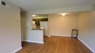 This single-floor space offers an open, functional layout connecting the kitchen and living area seamlessly. The flooring throughout is warm-toned hardwood-style planks, lending a clean and cohesive aesthetic across the space.

The kitchen is positioned behind a raised peninsula counter, which provides a natural division between the cooking and living zones while maintaining an open feel. Dark wood cabinetry with glass-paneled upper doors lines the kitchen walls, complemented by a mosaic tile backsplash that adds a subtle decorative element. A white refrigerator anchors one end of the kitchen, and a sink with a classic faucet fixture is visible along the counter. Under-cabinet lighting illuminates the workspace efficiently.

The adjacent living area is generously proportioned, with neutral beige walls and white baseboards that create a bright, understated backdrop. Ceiling-mounted light fixtures provide ambient illumination throughout. The textured ceiling adds a modest architectural detail, while the overall layout offers flexibility for various furniture arrangements. The space presents a practical and tidy environment suitable for everyday living.