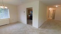 This single-floor residential unit offers a straightforward and functional layout designed for comfortable everyday living. The main living area is finished with wall-to-wall carpeting in a neutral tone, creating a warm and understated atmosphere. Clean, light-colored walls throughout contribute to a bright and open feel, keeping the space feeling airy despite its modest footprint.

A wide doorway connects the living area to the kitchen, providing an open sightline between the two spaces. The kitchen features tile flooring, warm-toned wooden upper and lower cabinetry, and black appliances including a gas range and over-the-range microwave. The layout is compact and efficient, making good use of the available space.

Electrical outlets and light switches are positioned throughout at standard heights, and the smooth ceilings are free of heavy ornamentation, keeping the design simple and clean. The overall ambiance of the unit is practical and neutral, offering a blank canvas well-suited for personalizing with furnishings and décor. The seamless flow between the living and kitchen areas enhances the sense of connectivity throughout the single-floor plan.