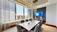 Located at 1909 Walnut St in Philadelphia's prestigious Rittenhouse Square neighborhood, this elegantly appointed conference room delivers a refined and professional environment designed for high-impact meetings. The space is anchored by a large marble-top conference table surrounded by plush upholstered chairs, offering comfortable seating for a full group of attendees. A dramatic sculptural chandelier with cascading gold and crystal elements serves as the room's defining design feature, suspended from a coffered ceiling with integrated ambient lighting that adds warmth and sophistication. Floor-to-ceiling windows line the exterior wall, flooding the room with abundant natural light and framing impressive views of the Philadelphia skyline and surrounding cityscape. Sheer drapery panels provide flexible light control without compromising the airy, open feel. A dedicated presentation wall features bold geometric wallcovering in deep navy tones, against which a large-format display screen is mounted and flanked by gold accent lamps, creating a polished focal point ideal for video conferencing and presentations. Sliding glass partitions with dark metal framing connect the space to adjacent areas, allowing for flexible configurations. Curated abstract artwork adds a creative touch, completing a space that balances functionality with upscale design.