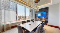 Located at 1909 Walnut St in Philadelphia's prestigious Rittenhouse Square neighborhood, this elegantly appointed conference room delivers a refined and professional environment designed for high-impact meetings. The space is anchored by a large marble-top conference table surrounded by plush upholstered chairs, offering comfortable seating for a full group of attendees. A dramatic sculptural chandelier with cascading gold and crystal elements serves as the room's defining design feature, suspended from a coffered ceiling with integrated ambient lighting that adds warmth and sophistication. Floor-to-ceiling windows line the exterior wall, flooding the room with abundant natural light and framing impressive views of the Philadelphia skyline and surrounding cityscape. Sheer drapery panels provide flexible light control without compromising the airy, open feel. A dedicated presentation wall features bold geometric wallcovering in deep navy tones, against which a large-format display screen is mounted and flanked by gold accent lamps, creating a polished focal point ideal for video conferencing and presentations. Sliding glass partitions with dark metal framing connect the space to adjacent areas, allowing for flexible configurations. Curated abstract artwork adds a creative touch, completing a space that balances functionality with upscale design.