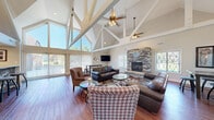 This single-floor space makes an immediate architectural statement with its dramatic vaulted ceilings supported by exposed white-painted timber trusses that span the full width of the main gathering area. Dual ceiling fans are suspended from the peak, complementing the abundance of natural light that pours in through floor-to-ceiling windows and decorative leaded glass panes along the walls.

The open-concept main area seamlessly integrates a spacious living room and a casual bar-height dining area. The living room is anchored by a striking floor-to-ceiling stacked stone fireplace, which serves as the focal point of the room and is framed by warm hardwood flooring that runs throughout. Leather sofas and houndstooth-patterned accent chairs provide comfortable, stylish seating arranged around a glass-top coffee table.

The kitchen is efficiently designed with stainless steel appliances — including a range, refrigerator, and microwave — set below recessed lighting. A substantial stone-clad breakfast bar with a granite-style countertop separates the kitchen from the main living space, offering additional casual seating.

A transitional entry area features a decorative stacked stone accent wall, a frosted-glass interior door, and a checkered tile floor, leading to a separate carpeted room that appears suited for office or service use. The cohesive use of stone, wood, and white structural elements gives the entire space a warm yet architecturally refined character.