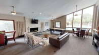 This single-story clubhouse offers a thoughtfully designed gathering space that blends comfort and recreation seamlessly. The main lounge area is expansive and open, anchored by a striking floor-to-ceiling white stone fireplace with a wall-mounted flat-screen TV above it. Decorative birch branch arrangements flank the fireplace, adding a natural, organic touch to the space. Comfortable seating, including a tufted leather sofa and patterned accent chairs arranged over bold area rugs, creates an inviting atmosphere for socializing and relaxation. A pool table positioned near large floor-to-ceiling windows allows natural light to flood the room while offering views of the outdoor pool area beyond. Ceiling fans are installed throughout to maintain comfort. A dedicated dining nook with table and chairs provides a casual spot for meals or meetings. The clubhouse also includes a functional kitchen equipped with dark granite countertops, wood cabinetry, black appliances including a refrigerator, range, and microwave, along with a sink and coffee station. The overall ambiance is warm and contemporary, with a neutral color palette, varied textures, and a layout that accommodates both quiet gatherings and lively group activities with ease.