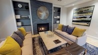 This thoughtfully designed lobby offers a warm and inviting atmosphere that immediately sets a sophisticated tone. The space is anchored by a striking lounge area centered around a built-in fireplace clad in stacked dark slate tile, flanked by recessed shelving with cabinetry in a cool slate-blue finish. A bold sunburst wall sculpture in brushed gold crowns the fireplace, serving as a dramatic focal point. Plush neutral sofas adorned with vivid mustard yellow and navy geometric throw pillows surround a herringbone-patterned coffee table atop a richly textured area rug, creating an intimate and stylish seating arrangement.

The main lobby area opens to a spacious, light-filled room with soaring ceilings and large floor-to-ceiling windows dressed in soft linen drapes, allowing abundant natural light to flood the space and offering views of the exterior courtyard. Light wood-look tile flooring flows throughout, lending a clean, contemporary feel underfoot. A cluster of bold cobalt blue wingback chairs arranged around a round accent table provides an additional seating vignette beneath an elegant chandelier. Abstract artwork punctuates the walls with pops of yellow and blue, tying the color palette together. The overall ambiance is refined yet approachable, blending comfort with contemporary design.