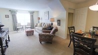 This single-floor residence offers a thoughtfully arranged living space with a warm, traditional aesthetic. The living room serves as the focal point, featuring plush wall-to-wall carpeting in a neutral beige tone that adds comfort underfoot and contributes to the space's inviting atmosphere. The room accommodates a classic sofa paired with a richly patterned armchair and a coordinating upholstered ottoman, creating a cozy and functional seating arrangement. A textured accent wall and a decorative art piece add visual interest, while table lamps provide warm ambient lighting throughout. A window dressed with elegant valance curtains allows natural light to filter in, brightening the space. The angled ceiling detail adds an architectural element that gives the room character and dimension. To one side, a dining area with dark wood chairs is partially visible, suggesting a seamless transition between living and dining spaces. A doorway leads further into the residence, hinting at additional rooms beyond. The overall ambiance is calm, classic, and residential, well-suited for comfortable everyday living.