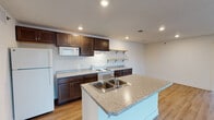 This single-floor residence at 3233 Lake Michigan Dr NW in Grand Rapids, Michigan offers a well-organized layout with distinct living, kitchen, bedroom, and bathroom areas. The open-concept living space is anchored by warm hardwood-style flooring that extends throughout the main area, with a sliding glass door at one end drawing in natural light and providing access to an outdoor space.

The kitchen is a functional centerpiece of the unit, featuring dark espresso cabinetry with brushed nickel hardware paired with granite-look countertops. A central island with a double basin sink adds workspace and visual interest. White appliances, including a refrigerator, range, and microwave, complement the design, while open wood shelving provides additional storage and display space.

The bedrooms offer neutral carpet flooring and clean white walls, creating a calm and versatile environment. Two bathrooms serve the unit, each finished with hexagonal mosaic tile flooring and dark wood vanities with light countertops. One bathroom features a tub-shower combination, while the other includes a standalone shower with built-in shelving and adjacent linen storage. A dedicated laundry room houses a full-size washer and dryer, adding everyday convenience.