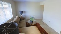 This single-floor space presents a straightforward and functional layout, centered around a modest living area finished in soft, neutral tones throughout. The walls are painted in a warm off-white, contributing to a clean and understated aesthetic that allows furnishings to take center stage. The flooring consists of light-colored carpeting, adding a sense of warmth underfoot across the main living zone. The living area is furnished with a generously sized sectional sofa in a muted tan upholstery, complemented by a round glass-top coffee table resting on a richly colored geometric area rug in warm reds and earthy tones, which anchors the seating arrangement. A classic table lamp with a dark shade adds ambient lighting beside the sofa. A small potted plant with blue flowers on the coffee table introduces a touch of natural color to the otherwise neutral palette. A recessed nook is visible along one wall, offering potential for additional storage or display use. The overall ambiance is calm and livable, with a simple, no-frills character suited to comfortable everyday use.
