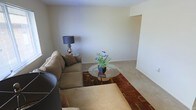 This single-floor space presents a straightforward and functional layout, centered around a modest living area finished in soft, neutral tones throughout. The walls are painted in a warm off-white, contributing to a clean and understated aesthetic that allows furnishings to take center stage. The flooring consists of light-colored carpeting, adding a sense of warmth underfoot across the main living zone. The living area is furnished with a generously sized sectional sofa in a muted tan upholstery, complemented by a round glass-top coffee table resting on a richly colored geometric area rug in warm reds and earthy tones, which anchors the seating arrangement. A classic table lamp with a dark shade adds ambient lighting beside the sofa. A small potted plant with blue flowers on the coffee table introduces a touch of natural color to the otherwise neutral palette. A recessed nook is visible along one wall, offering potential for additional storage or display use. The overall ambiance is calm and livable, with a simple, no-frills character suited to comfortable everyday use.