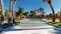 This expansive outdoor amenity area showcases a beautifully designed resort-style environment ideal for relaxation and recreation. At its centerpiece is a generously sized freeform swimming pool with smooth, curved edges and a shallow entry, finished in light-toned tile that reflects the brilliant blue sky above. The pool deck is finished in a textured concrete surface, providing ample space for lounging, and is outfitted with modern sling-style chaise loungers and oversized shade umbrellas positioned throughout.

Surrounding the pool, towering mature palm trees and lush deciduous trees provide natural shade and a tropical ambiance, enhancing the desert-resort aesthetic. The grounds are meticulously landscaped with manicured green turf areas that contrast beautifully against the warm earth tones of the surrounding multi-story stucco buildings, which feature tiled rooftops and private balconies.

A dedicated lounge and social area is anchored by a stone fire pit surrounded by blue cushioned chairs, creating an inviting gathering space. Pergola-style structures with brick columns and metal gates add architectural character and define separate zones within the courtyard. Blue-draped cabana structures line portions of the perimeter, adding a boutique, upscale touch. The entire space enjoys abundant natural sunlight under consistently clear skies, making it a vibrant, well-appointed outdoor retreat.