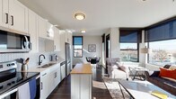 This contemporary single-floor unit offers a thoughtfully designed open-concept layout that seamlessly connects the kitchen, dining, and living areas. The kitchen is a standout feature, outfitted with crisp white shaker cabinetry accented by matte black hardware, a herringbone-patterned tile backsplash, stainless steel appliances, and a butcher block island that serves as both a prep space and casual gathering spot. Rich, dark hardwood flooring runs throughout the main living areas, grounding the bright, neutral palette.

The living room is bathed in natural light through expansive floor-to-ceiling windows dressed with sleek roller shades, framing breathtaking elevated views of the city below and a mountain range in the distance. A compact dining area sits adjacent, completing the open social space.

Two well-proportioned bedrooms continue the modern aesthetic with plush carpeting, large windows offering sweeping city views, and clean white walls that create a calm, restful atmosphere. The bathroom features large-format tile flooring, white cabinetry with black hardware, and a tub-shower combination. Throughout the unit, consistent use of black-framed windows and fixtures ties the design together with a refined, contemporary edge.