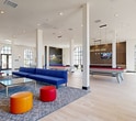 Located at 1900 E Inland Empire Blvd in Ontario, California, this single-floor community amenity space offers a generously sized, open-plan layout designed for recreation and socializing. The interior is defined by soaring ceilings supported by white structural columns, and an abundance of natural light streaming through dramatic arched, floor-to-ceiling windows with black steel frames that line the perimeter walls.

Light hardwood flooring extends throughout the entire space, lending a clean, contemporary aesthetic. The main game and recreation area features multiple billiard tables with vibrant red felt, a ping-pong table, foosball, and classic arcade machines — all thoughtfully arranged to maximize flow and usability. A dedicated lounge zone anchors the center of the space with a bold blue sectional sofa, colorful ottomans, and a glass coffee table atop a patterned area rug, complemented by a wall-mounted flat-screen TV.

A standout wet bar area showcases a dramatic dark marble waterfall-edge island, wood cabinetry, glossy textured tile backsplash, and elegant pendant lighting — ideal for hosting events. Dining tables with modern chairs are positioned near the windowed walls to take advantage of the outdoor views. Recessed lighting and pendant fixtures provide warm, even illumination throughout, completing this vibrant, well-appointed gathering space.