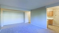 This single-floor residential space offers a clean, functional layout designed for comfortable everyday living. The interior is characterized by soft, neutral gray-toned walls that create a calm and understated ambiance throughout. The living area is finished with plush wall-to-wall carpeting in a muted blue-gray hue, providing a cozy underfoot feel while contributing to the overall soft color palette of the space. Bright white baseboards and trim provide crisp contrast against the walls, lending a tidy and well-maintained appearance. The ceiling is smooth and painted white, helping to reflect light and give the room an open, airy feel. A partial wall or structural beam element adds a subtle architectural detail, subtly defining areas within the open floor plan. A white interior door leads to an adjoining area, suggesting a connected layout between rooms. Electrical outlets are conveniently placed along the walls, and a ceiling vent indicates a central climate control system. The overall atmosphere is straightforward and move-in ready, offering a neutral canvas well-suited for a variety of interior styles and personal touches.