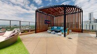 Located atop 848 Spring St NW in the heart of Atlanta, GA, this rooftop amenity deck offers a stunning urban retreat with sweeping panoramic views of the Atlanta skyline. The space is thoughtfully designed to accommodate a variety of outdoor experiences, combining multiple distinct zones across its open-air layout.

A welcoming entry with floor-to-ceiling glass doors and brick accents provides immediate access to the terrace, where the city skyline unfolds as a dramatic backdrop. One lounge area features a cozy outdoor fireplace flanked by plush, blue-cushioned sectional sofas and is framed by a wall-mounted TV — ideal for relaxed gatherings. A lush turf lawn area adds a touch of greenery and a casual space for recreation.

A standout architectural feature is the curved wooden pergola, outfitted with string lights and vibrant teal seating, creating an intimate social nook with a stylish neon sign as a focal point. Adjacent to this, a covered grilling station with multiple BBQ units sits beneath a slatted wood canopy, accompanied by picnic-style communal dining tables and eclectic modern seating. The perimeter of the deck features glass-panel railings throughout, ensuring unobstructed views of the Atlanta cityscape from every angle. The overall ambiance is energetic yet inviting, blending modern urban design with functional outdoor living.