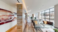 This contemporary single-floor lobby space is defined by its sophisticated blend of materials, natural light, and thoughtfully arranged seating zones. Floor-to-ceiling curtain-wall windows line the perimeter, flooding the interior with daylight and offering unobstructed views of the surrounding outdoor landscape. The polished marble-effect flooring reflects ambient light throughout, enhancing the open, airy atmosphere.

The layout is organized into distinct yet connected lounge areas. One zone features sleek teal leather sofas paired with accent chairs and a low coffee table atop a patterned area rug, ideal for casual gatherings. An adjacent seating area offers warm tan velvet sofas arranged around low tables, complemented by wall-mounted artwork and a bar-height counter for individual work. A rounded corner nook contains a circular high-top table surrounded by mixed seating, well-positioned to capture natural light from two window walls.

Architectural detailing is a highlight throughout, with a wood-clad structural column featuring integrated uplighting, cove-lit ceiling trays, and curated artwork. A separate corridor leads to restroom facilities, finished with dark marble-tiled walls, hardwood flooring, and a built-in water station. The overall ambiance is polished, welcoming, and conducive to both collaboration and focused work.