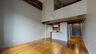 This two-floor loft-style space masterfully combines industrial heritage with contemporary updates. The main level opens into a generous living area defined by dramatically soaring ceilings and prominent exposed wood beams that lend a sense of warmth and raw character. Rich, honey-toned hardwood floors extend throughout, complementing the open layout and neutral gray walls. A peninsula-style kitchen is tucked neatly to one side, featuring dark wood cabinetry, granite countertops, stainless steel appliances including a built-in microwave and range, and overhead task lighting — all continuing the consistent beam detail above. A dedicated laundry area with a stacked washer and dryer adds practical convenience. The bathroom is finished with a granite-topped dark wood vanity, large-format white subway tile walk-in shower, and neutral floor tile. The upper level offers a carpeted loft space with a ceiling fan, open railing overlooking the main floor below, and access to an additional room framed by the same signature exposed beams. Large multi-pane windows on the upper level flood the space with natural light. Together, the two floors deliver a cohesive, industrial-chic aesthetic with thoughtful modern amenities throughout.