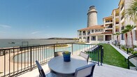 This remarkable waterfront property is defined by its striking architectural character and exceptional outdoor amenities. The centerpiece of the exterior is a tall, cylindrical stone lighthouse that rises prominently above the complex, lending a iconic coastal identity to the building. The main structure is constructed of light-toned limestone with arched windows, covered colonnades, and a terracotta-tiled roofline, blending Mediterranean and coastal Texas architectural influences seamlessly.

The outdoor living space is a standout feature, anchored by a generously sized, freeform swimming pool with crystal-clear water and a wide, curved concrete surround. The pool deck is outfitted with modern lounge chairs and side tables set atop manicured artificial turf, creating comfortable areas for relaxation. Mature palm trees are strategically placed throughout the grounds, reinforcing the resort-like atmosphere.

Elevated terraces and private balconies extend from the building's upper levels, several of which are furnished with outdoor dining sets and offer sweeping, unobstructed views of the adjacent waterway and a long bridge stretching to the horizon. The entire property is oriented toward the water, making panoramic open-water views a defining characteristic of the experience. The combination of thoughtful landscaping, quality stonework, and waterfront positioning creates a distinctive and serene environment.