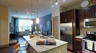 This single-floor space showcases a thoughtfully designed open-concept layout that seamlessly connects the kitchen and living areas. The kitchen is a standout feature, anchored by a generous island topped with elegant granite countertops that provide ample workspace and casual seating. Dark espresso cabinetry offers a rich contrast to the light stone surfaces, while a full suite of stainless steel appliances, including a French-door refrigerator, adds a polished, modern touch. A combination of pendant lights and track lighting illuminates the space with both style and practicality, casting a warm glow throughout. The open layout transitions naturally into the living and dining areas, where large windows invite an abundance of natural light and offer views of the surrounding greenery. The living space is finished with neutral tones and contemporary furnishings, creating a relaxed yet sophisticated ambiance. Consistent flooring flows throughout, visually unifying the entire space. The overall design strikes a balance between modern aesthetics and everyday functionality, making it an inviting and well-appointed environment.
