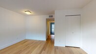 This single-floor unit is designed with a clean, modern aesthetic and consistent light hardwood flooring that runs throughout the main living areas and bedroom, creating a warm, cohesive feel. The living area is bright and open, featuring white walls, a large window with horizontal blinds that allows natural light to filter in, and a gray front door that provides direct access to the exterior. A hallway connects the main living space to the bedroom, which offers a closet and a flush-mount ceiling light fixture, maintaining the same minimalist character found throughout the unit.

The kitchen is outfitted with stainless steel appliances, including an electric range and an over-the-range microwave, complemented by dark granite countertops, warm wood-toned cabinetry, and a charcoal subway tile backsplash that adds a modern contrast. The bathroom features dark diagonal floor tiles, a full soaking tub, and floor-to-ceiling white basketweave tile surround, contributing a clean and timeless design. HVAC vents are visible in the ceiling, ensuring climate control throughout. Overall, the space presents a practical yet stylish interior with consistent finishes and a neutral palette suited to a variety of uses.