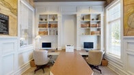 Located along Avemore Lane in Charlottesville, Virginia, this single-floor business center combines classic architectural detailing with fully equipped professional workspaces. The office area is defined by a symmetrical arrangement of built-in white shelving units framed by raised panel millwork that extends to the ceiling, lending the space a distinguished, library-like character. Multiple workstations are outfitted with desktop computers and ergonomic seating, supported by a shared printer, making the area well-suited for productivity. The shelving displays decorative books and curated accessories, adding a polished, collegiate aesthetic consistent with the Charlottesville setting.

A separate conference or meeting room features a rich burl wood table surrounded by upholstered chairs, illuminated by a brass chandelier overhead. Tall windows fitted with blinds allow natural light to filter in while maintaining privacy, and framed artwork on the soft blue-gray walls contributes to a calm, professional atmosphere. A built-in credenza and beverage station complement the room's functionality. Direct exterior door access provides convenient entry. Throughout the space, wainscoting, panel molding, and a large decorative antique map mural reinforce a cohesive, traditional design sensibility.