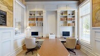 Located along Avemore Lane in Charlottesville, Virginia, this single-floor business center combines classic architectural detailing with fully equipped professional workspaces. The office area is defined by a symmetrical arrangement of built-in white shelving units framed by raised panel millwork that extends to the ceiling, lending the space a distinguished, library-like character. Multiple workstations are outfitted with desktop computers and ergonomic seating, supported by a shared printer, making the area well-suited for productivity. The shelving displays decorative books and curated accessories, adding a polished, collegiate aesthetic consistent with the Charlottesville setting.

A separate conference or meeting room features a rich burl wood table surrounded by upholstered chairs, illuminated by a brass chandelier overhead. Tall windows fitted with blinds allow natural light to filter in while maintaining privacy, and framed artwork on the soft blue-gray walls contributes to a calm, professional atmosphere. A built-in credenza and beverage station complement the room's functionality. Direct exterior door access provides convenient entry. Throughout the space, wainscoting, panel molding, and a large decorative antique map mural reinforce a cohesive, traditional design sensibility.