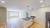 This thoughtfully designed single-floor residence offers a bright and contemporary living experience. The open-concept layout seamlessly connects the kitchen and living area, creating a spacious and functional environment. The kitchen serves as the centerpiece of the space, showcasing a large island with a dark granite countertop, an undermount sink, and pendant lighting above. White shaker-style cabinetry with matte black hardware lines the walls, complemented by matching dark granite counters. A full suite of stainless steel appliances — including a refrigerator, electric range, built-in microwave, and dishwasher — completes the well-equipped kitchen. Light hardwood flooring extends throughout the entire unit, lending warmth and continuity to each room. The living area benefits from natural light through large windows with outdoor views. Bedrooms are generously sized, with one featuring a walk-in closet and a ductless mini-split climate control unit. The bathroom features a white vanity with dark granite countertop, an undermount sink with a modern faucet, and a framed black mirror. Recessed lighting and neutral-toned walls throughout contribute to a clean, move-in-ready aesthetic.